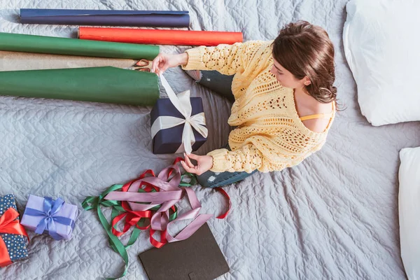 Blick von oben auf junge Frau, die Geschenkschachtel mit festlichem Band umwickelt, während sie zu Hause auf dem Bett sitzt — Stockfoto