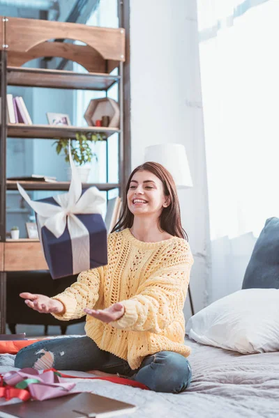 Selektiver Fokus des lachenden hübschen Mädchens, das Geschenkbox hochwirft, während es zu Hause auf dem Bett sitzt — Stockfoto