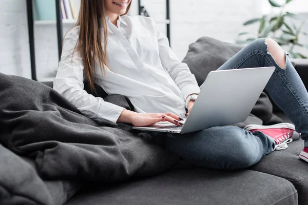 Обрезанный вид девушки, сидящей на диване и с помощью ноутбука в гостиной — стоковое фото