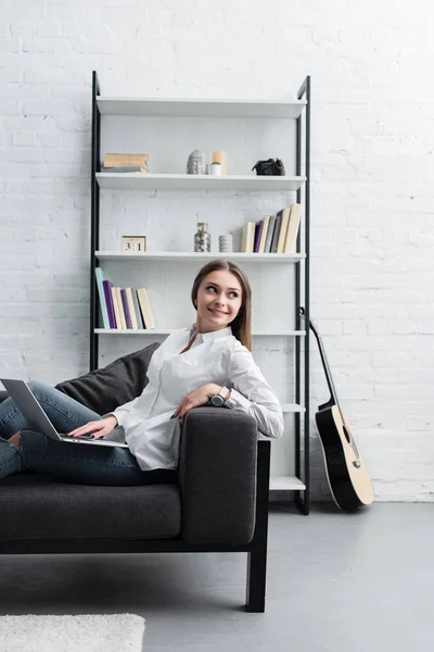 Красивая улыбающаяся девушка сидит на диване и с помощью ноутбука в гостиной — стоковое фото