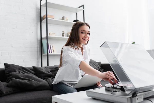Chica sonriente en camisa blanca sentado en el sofá con el ordenador portátil y tocando disco de vinilo en la sala de estar - foto de stock
