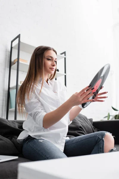 Beautiful girl in white shirt sitting on couch and holding vinyl record in living room — Stock Photo