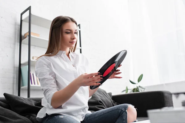 Attractive girl in white shirt sitting on couch and holding vinyl record in living room — Stock Photo