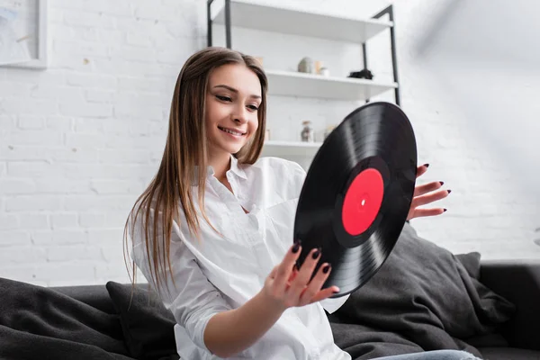 Sorridente ragazza in camicia bianca seduta sul divano e con record in vinile in salotto — Foto stock