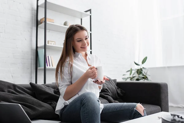 Усміхнена дівчина сидить на дивані і тримає чашку кави у вітальні — стокове фото