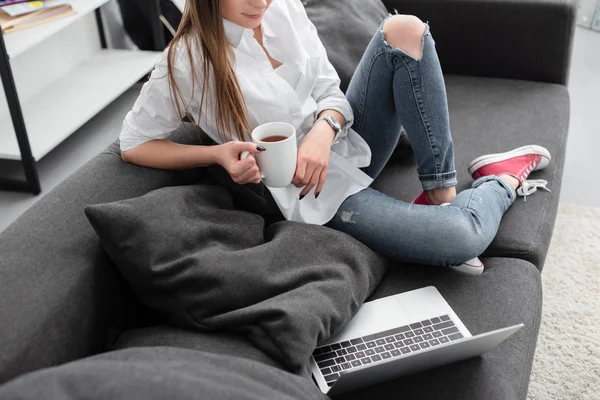 Partial view of girl holding coffee cup and sitting on couch with laptop in living room — Stock Photo