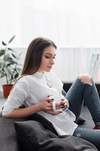 Chica pensativa sentada en el sofá y sosteniendo la taza de café en la sala de estar - foto de stock