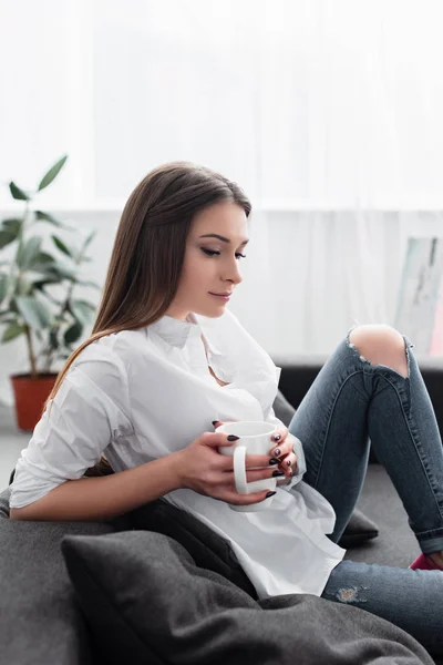 Menina pensativo sentado no sofá e segurando xícara de café na sala de estar — Fotografia de Stock
