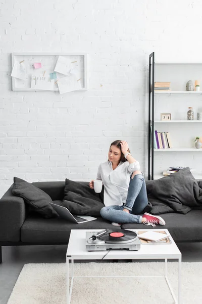 Chica sentada en el sofá con taza de café, el uso de ordenador portátil y escuchar reproductor de discos de vinilo en la sala de estar - foto de stock