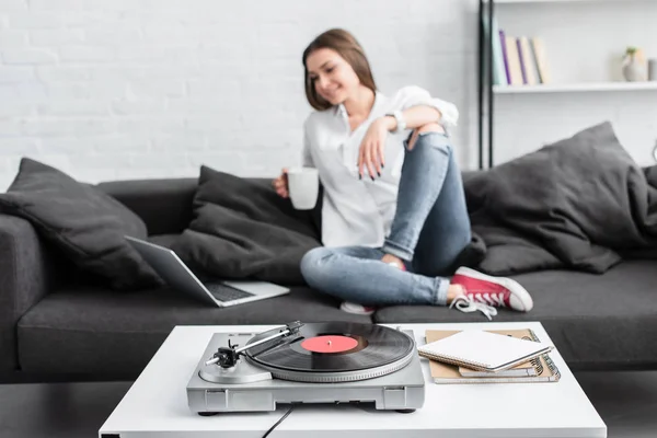 Mädchen in weißem Hemd sitzt auf Couch mit Kaffeetasse, benutzt Laptop und hört Plattenspieler im Wohnzimmer — Stockfoto