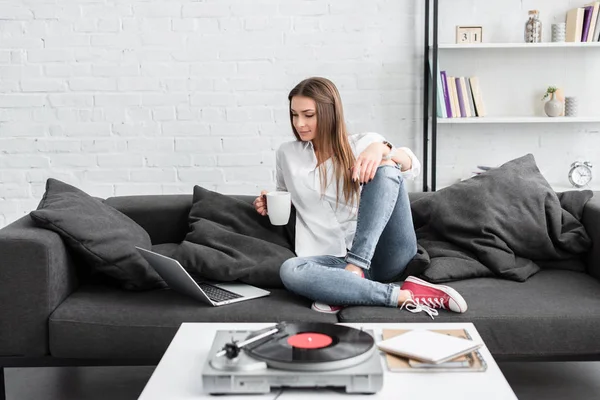 Belle fille en chemise blanche assise sur le canapé avec tasse de café, en utilisant un ordinateur portable et en écoutant un lecteur de disque vinyle dans le salon — Photo de stock