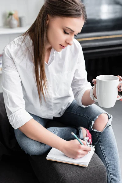 Attraente ragazza seduta con tazza di caffè e scrittura in notebook a casa — Foto stock