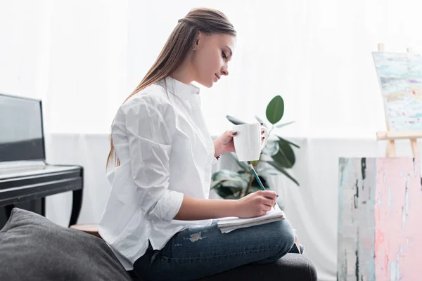 Mädchen im weißen Hemd sitzt mit Kaffeetasse auf Couch und schreibt in Notizbuch zu Hause — Stockfoto