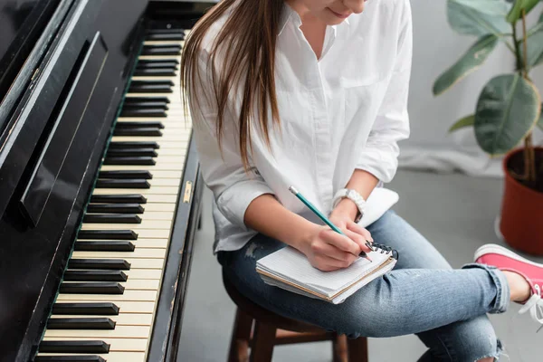 Обрезанный вид девушки с ноутбуком сидит рядом с фортепиано и сочинение музыки в гостиной — стоковое фото