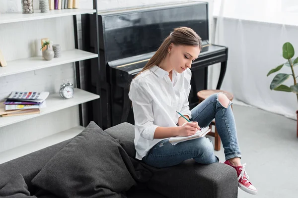 Mädchen mit Notizbuch sitzt auf Couch und komponiert Musik mit Klavier im Hintergrund im Wohnzimmer — Stockfoto