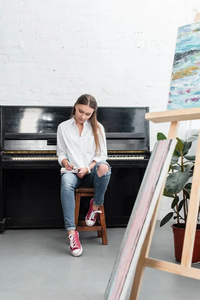 Fokussiertes Mädchen mit Notizbuch, das neben dem Klavier sitzt und im Wohnzimmer Musik komponiert — Stockfoto