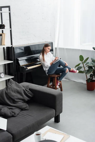 Chica con cuaderno sentado en la silla cerca del piano y componiendo música en la sala de estar - foto de stock