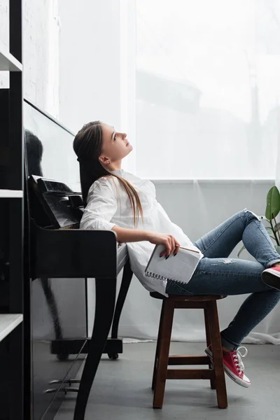 Menina sonhadora com notebook sentado perto de piano na sala de estar — Fotografia de Stock