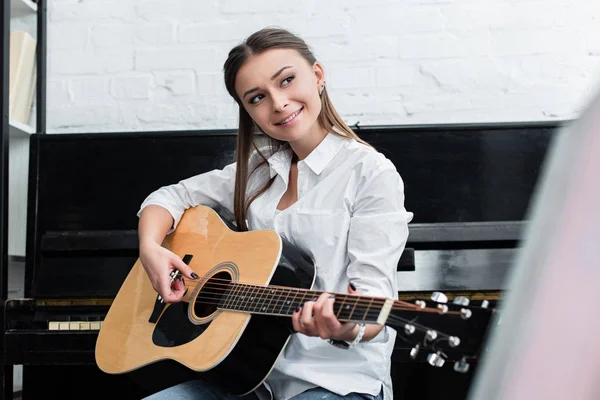 Menina sorrindo sentado e tocando guitarra na sala de estar com piano no fundo — Fotografia de Stock