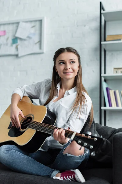 Hermosa chica sonriente sentado y tocando la guitarra en la sala de estar - foto de stock