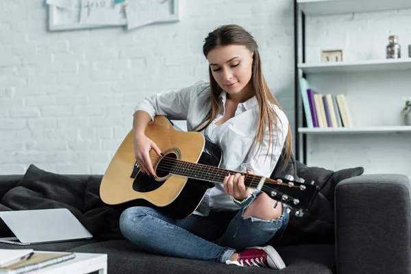 Fille concentrée assise sur le canapé et jouer de la guitare dans le salon — Photo de stock