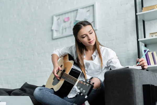 Красивая девушка сидит на диване с гитарой, пишет в блокноте и сочиняет музыку в гостиной — стоковое фото