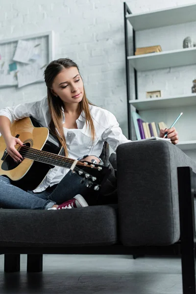 Задумчивая девушка сидит на диване с гитарой, пишет в блокноте и сочиняет музыку в гостиной — стоковое фото