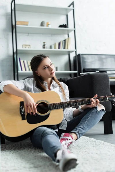 Bela menina pensativa sentado e tocando guitarra na sala de estar — Fotografia de Stock