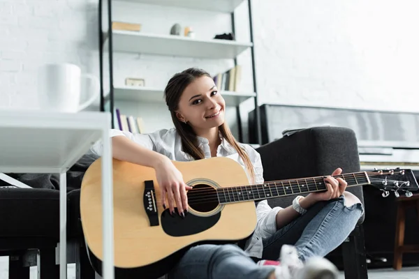 Hermosa chica sonriente sentado y tocando la guitarra en la sala de estar - foto de stock