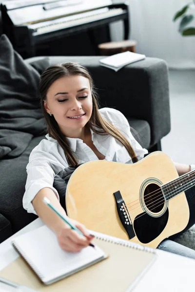 Fille souriante avec guitare acoustique écrit dans un carnet tout en composant de la musique à la maison — Photo de stock