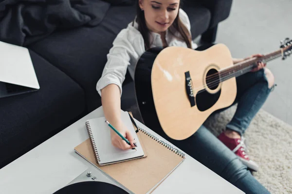 Mädchen mit Akustikgitarre sitzt auf dem Boden und schreibt in Notizbuch, während sie zu Hause Musik komponiert — Stockfoto