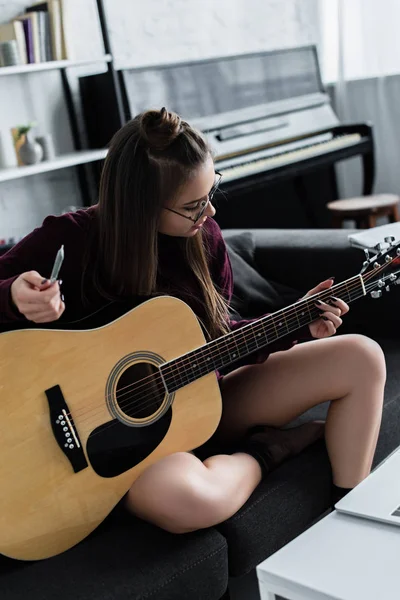Girl sitting on couch, playing guitar and holding marijuana joint in living room — Stock Photo