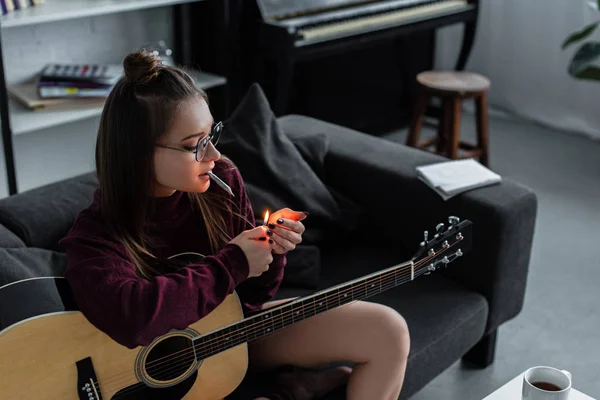Hermosa chica sentada, encendiendo marihuana conjunta y sosteniendo la guitarra en casa - foto de stock
