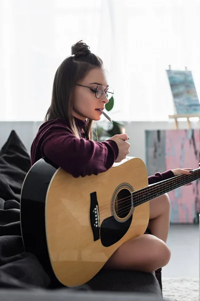 Привлекательная девушка сидит на диване, освещает марихуану и держит гитару дома — стоковое фото