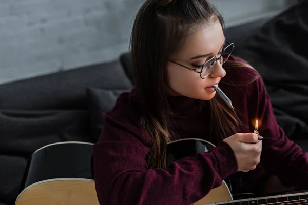 Fille dans des lunettes assis, éclairage joint de marijuana et tenant la guitare à la maison — Photo de stock