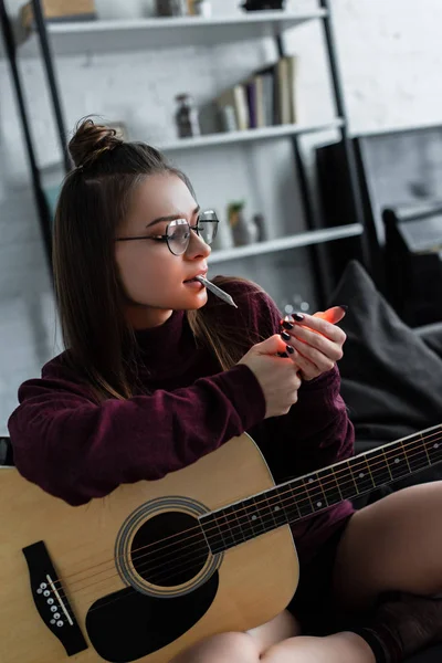 Belle fille assise sur le canapé, allumant joint de marijuana et tenant la guitare à la maison — Photo de stock