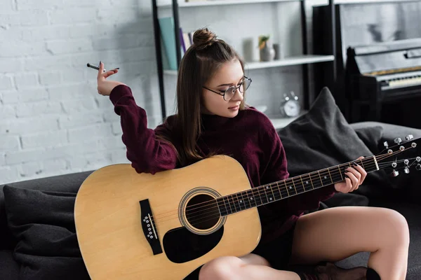 Chica atractiva sentada, sosteniendo marihuana conjunta y tocando la guitarra en casa - foto de stock
