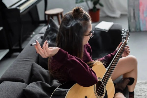 Girl sitting, holding marijuana joint and playing guitar in living room — Stock Photo