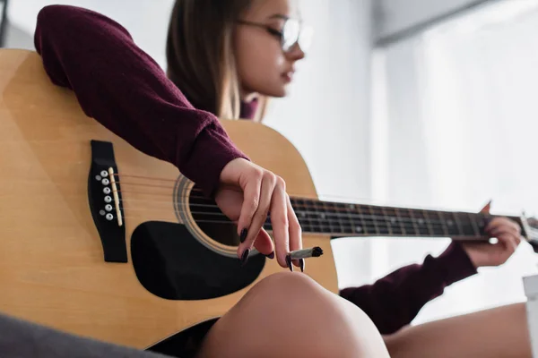 Избирательное внимание девушки, сидящей, держащей марихуану в руках и играющей дома на гитаре — стоковое фото
