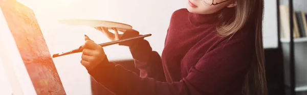 Cropped view of girl holding paintbrush with palette and painting at home with backlit — Stock Photo