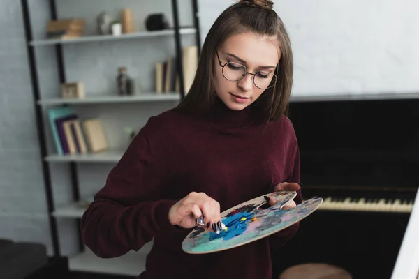 Girl in burgundy sweater with palette sitting and painting at home — Stock Photo