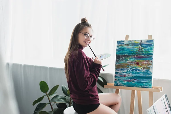 Smiling girl in glasses sitting in front of easel, holding paintbrush and palette and looking at camera in living room — Stock Photo