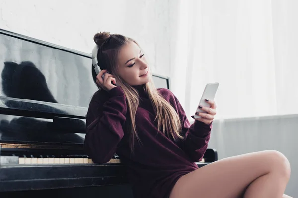 Привлекательная девушка с наушниками на голове сидя перед фортепиано и используя смартфон дома — Stock Photo