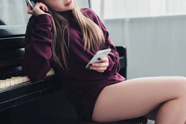 Vista cortada de menina com fones de ouvido na cabeça sentado na cadeira na frente do piano e segurando smartphone em casa — Fotografia de Stock