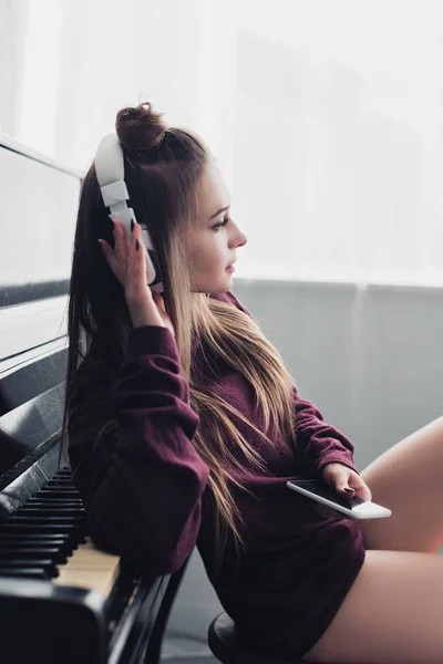 Привлекательная девушка с наушниками на голове сидит на стуле перед фортепиано и держит смартфон дома — стоковое фото
