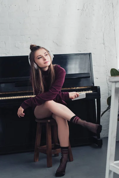 Menina bonita com fones de ouvido na cabeça sentado na cadeira na frente do piano e segurando smartphone na sala de estar — Fotografia de Stock