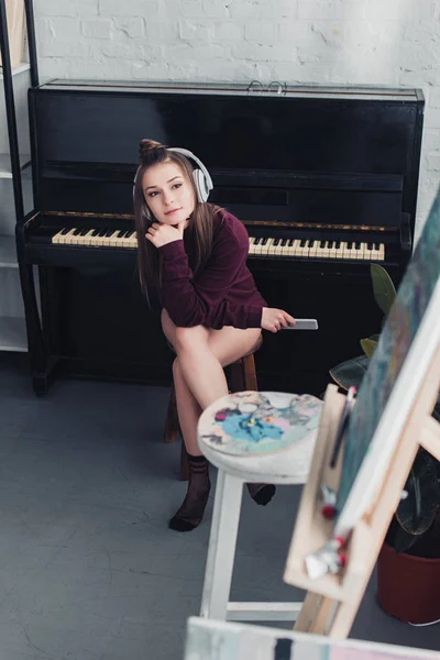 Beautiful girl with headphones on head sitting on chair in front of piano, holding smartphone and looking at easel in living room — Stock Photo