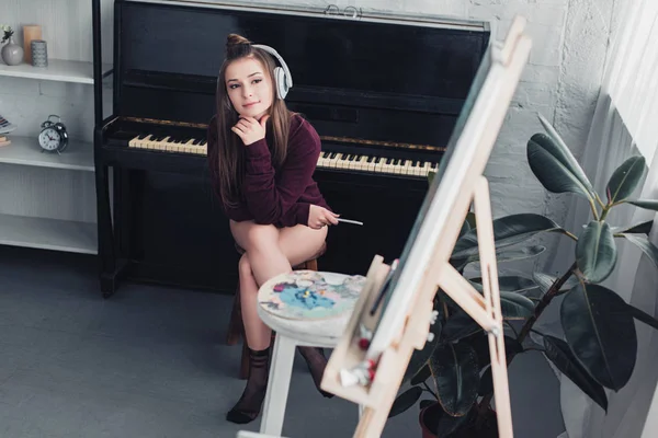 Attractive girl with headphones on head sitting on chair in front of piano, holding smartphone and looking at easel in living room — Stock Photo