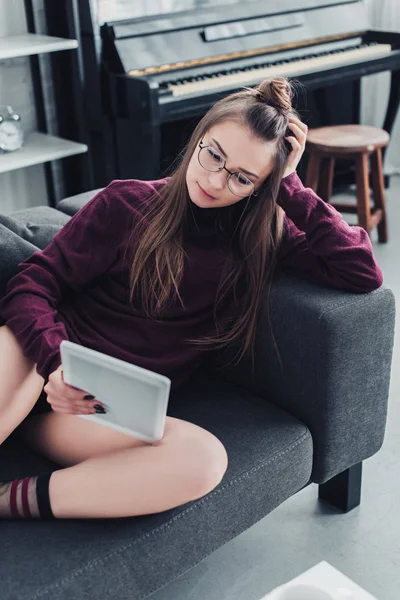 Schöne Mädchen mit Brille auf dem Sofa liegend und mit digitalem Tablet im Wohnzimmer — Stockfoto