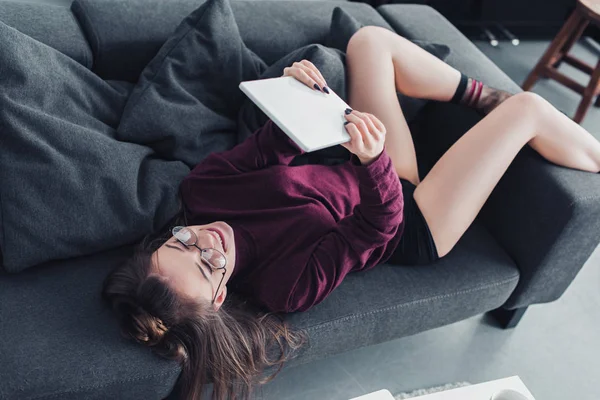 Chica sonriente tumbada en el sofá y utilizando la tableta digital - foto de stock